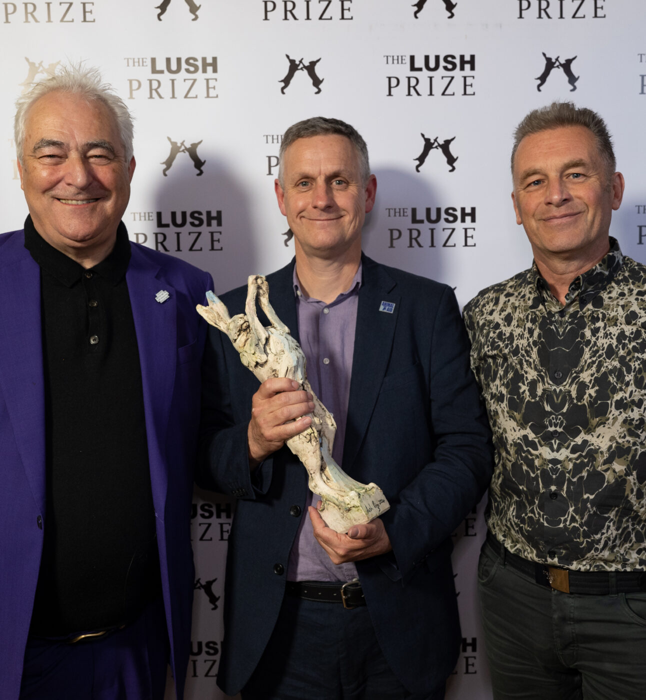 Three men pose smiling for a photo. Iain holds a trophy of a hare.