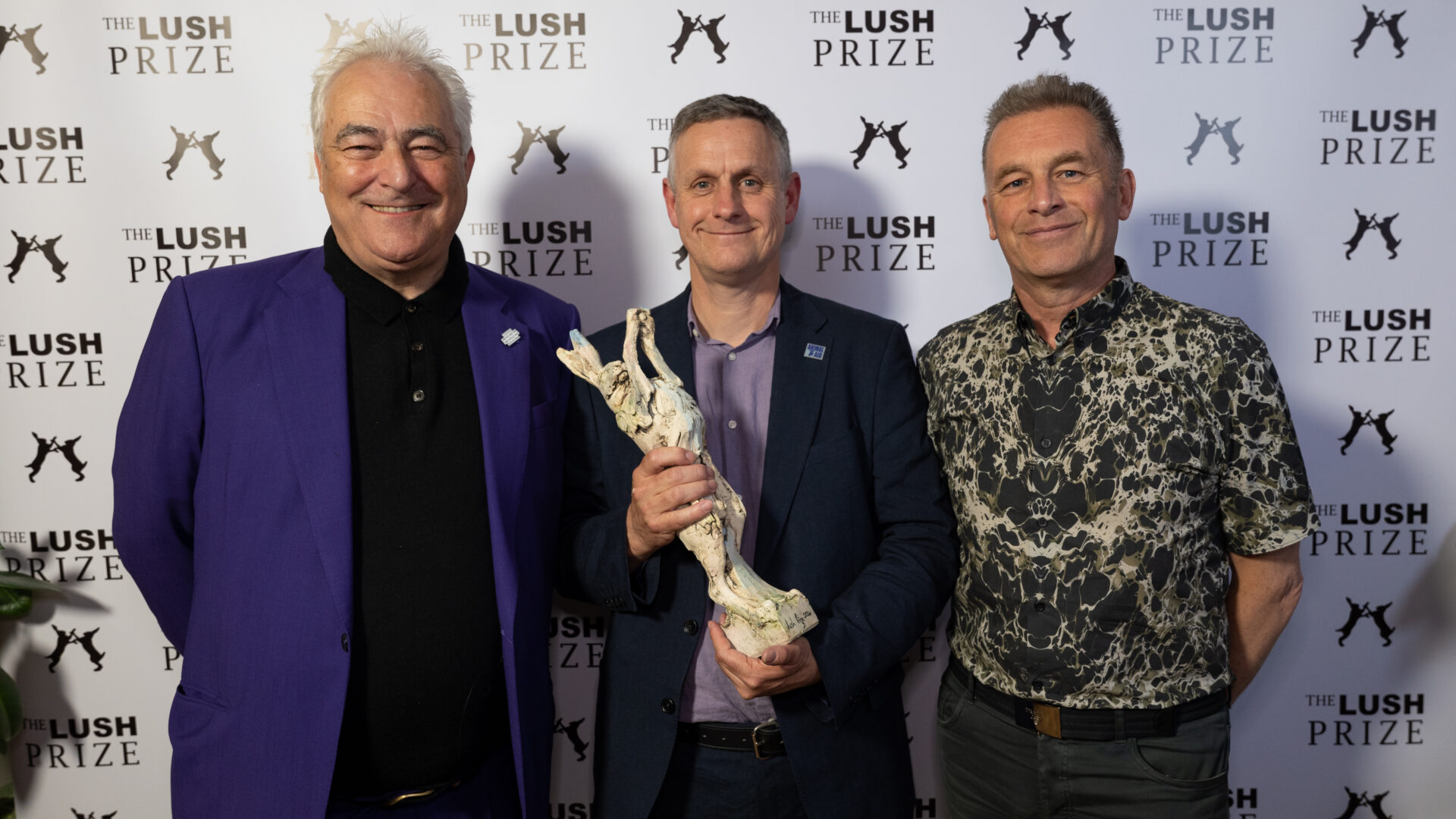 Three men pose smiling for a photo. Iain holds a trophy of a hare.