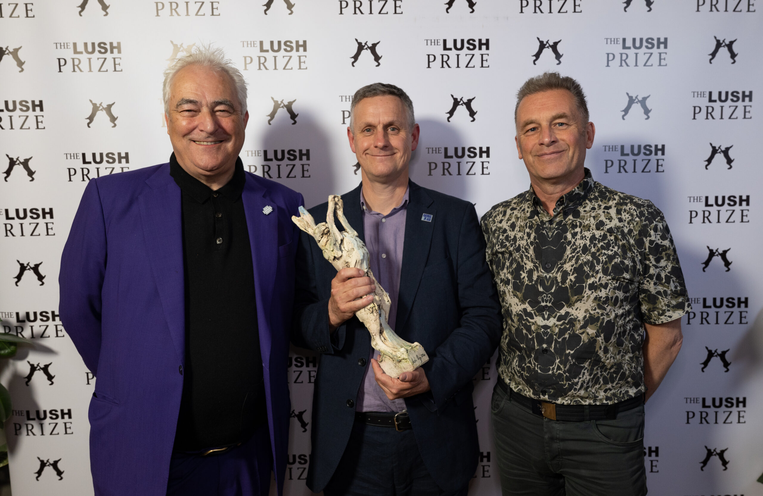 Three men pose smiling for a photo. Iain holds a trophy of a hare.